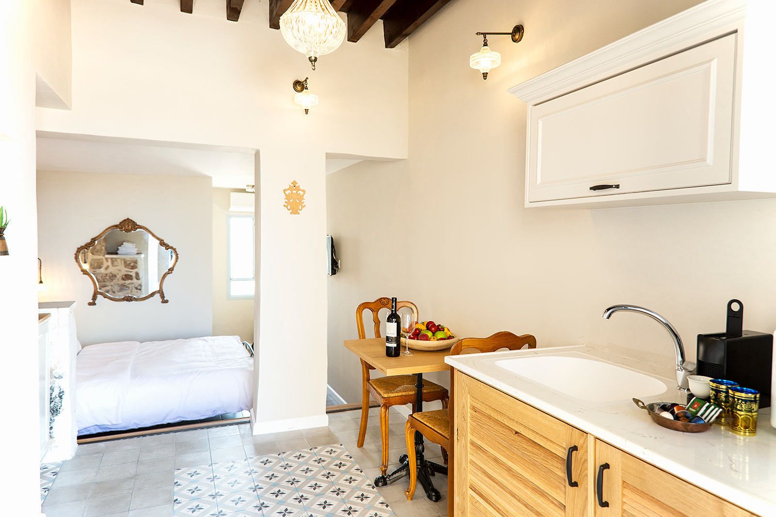 Habitación de hotel compacta con cama, una pequeña mesa de comedor con una botella de vino y fruta, un lavamanos con grifo moderno y un espejo decorativo.