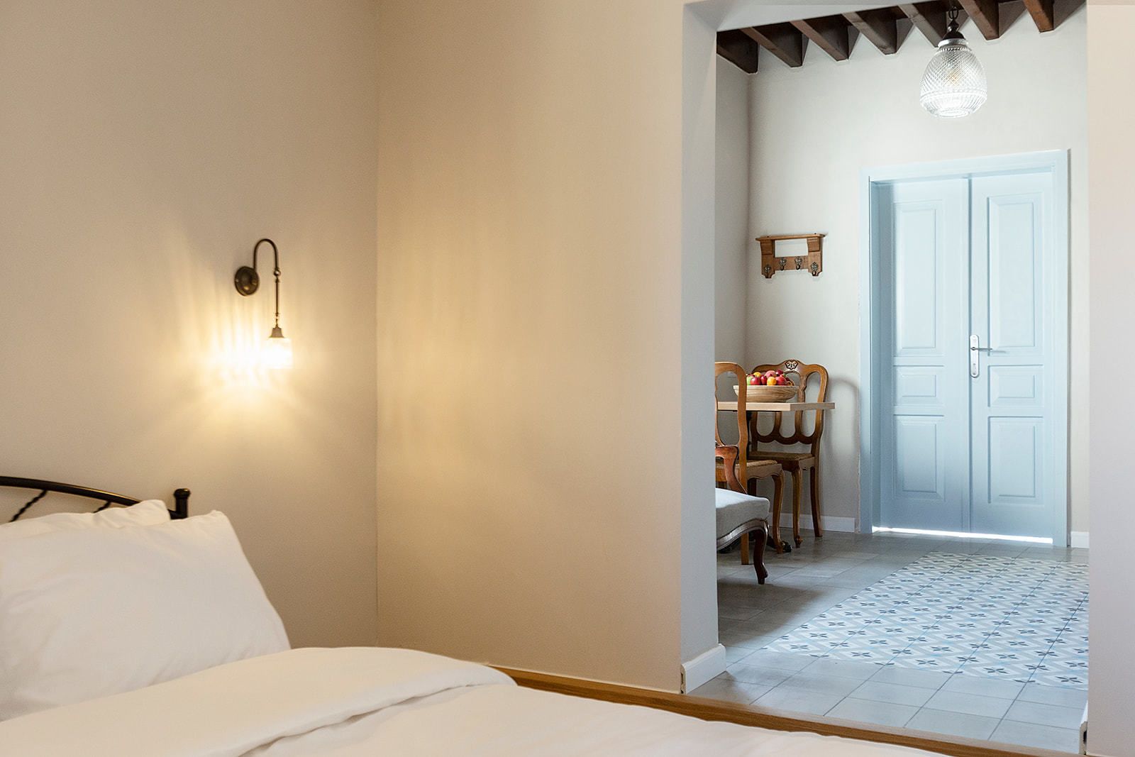A bedroom with a bed, a nightstand, a wall-mounted lamp, and a view into a dining area with a table and chairs.