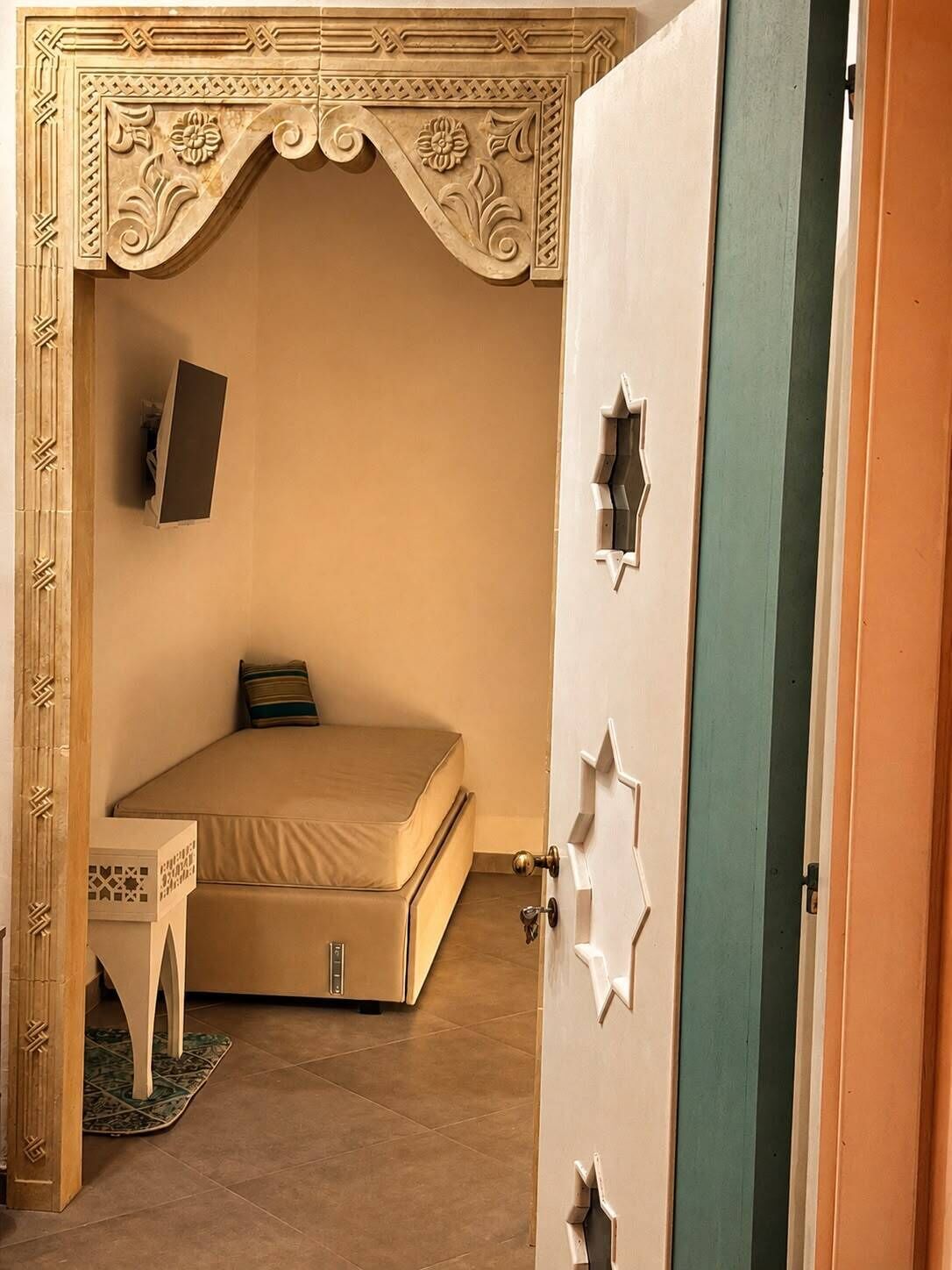 A room with a bed, a small table, a TV mounted on the wall, and a door with star-shaped cutouts.