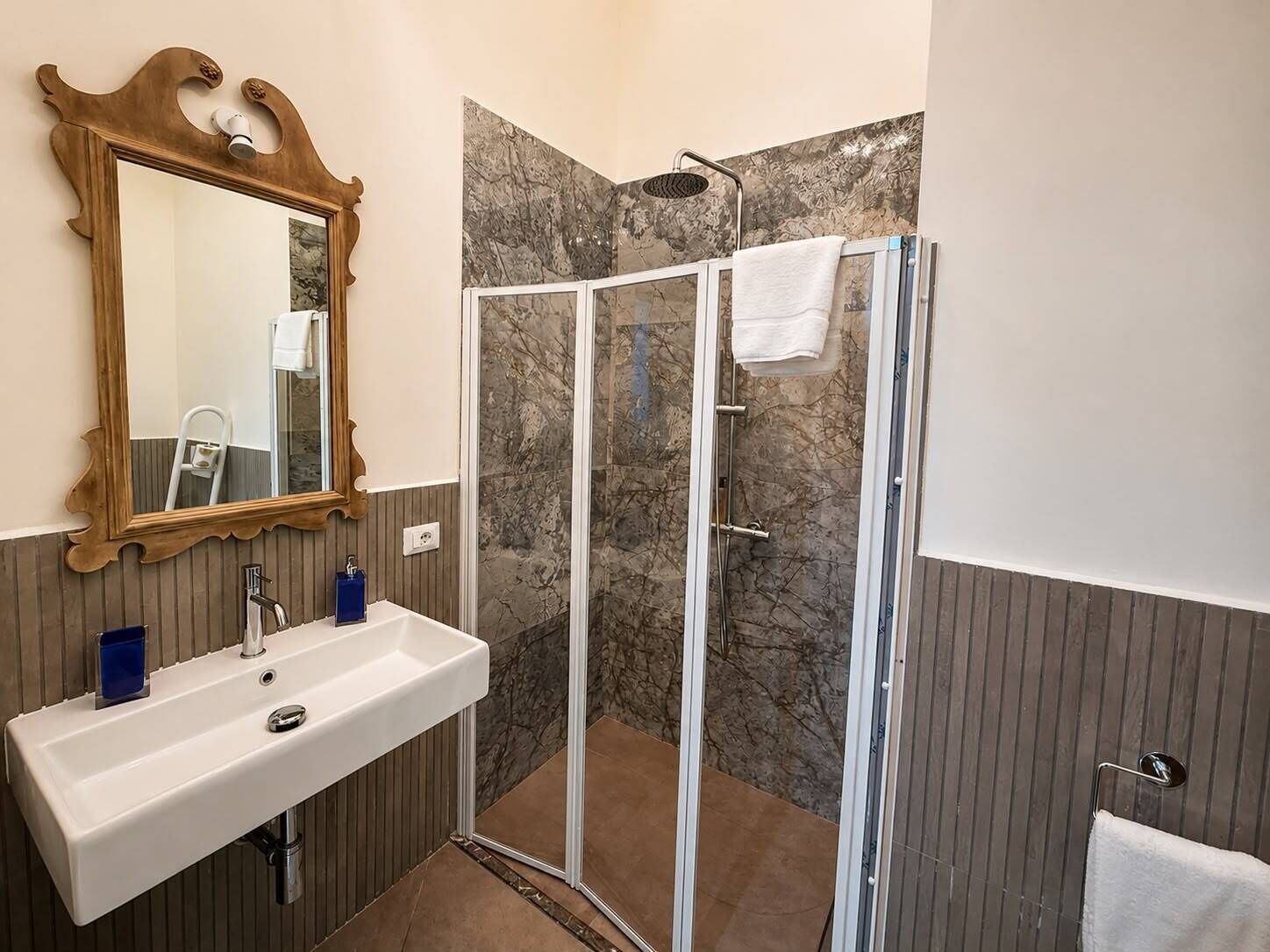 Ein Badezimmer mit einem weißen Waschbecken, einem Spiegel mit aufwendiger Rahmung, einer Dusche mit Glastür und einem Handtuchhalter.