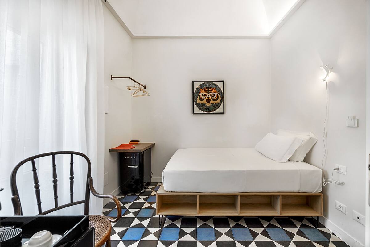 A small, neatly arranged bedroom with a bed, a chair, a desk, and a wall-mounted light fixture.