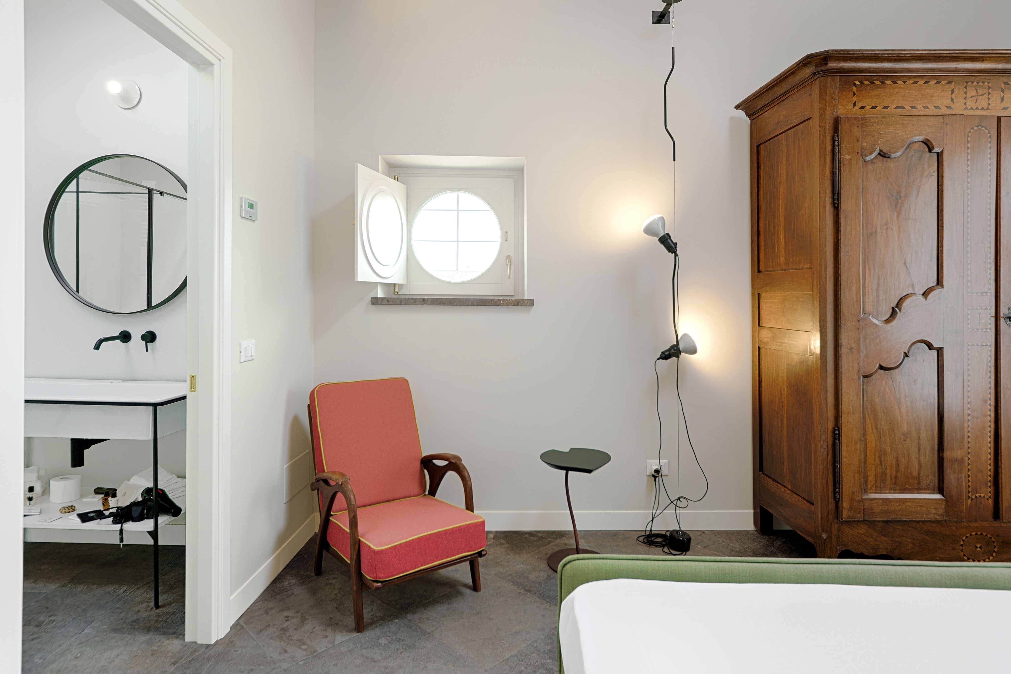 Een kamer met een bed, een houten kast, een stoel, een klein tafeltje, een spiegel en een raam met een lamp.