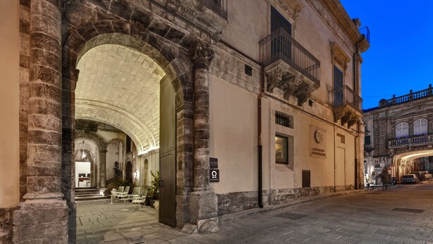 An old building with a large arched entrance, a sign indicating a historical site, and a street with a few parked cars. - a.d. 1768 Boutique Hotel (Ragusa)