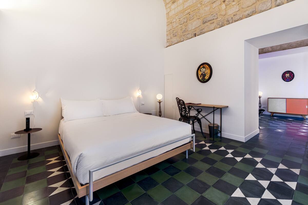 A bedroom with a bed, bedside tables, a desk, and a chair. There is a wall with a circular artwork and a stone wall above the bed.