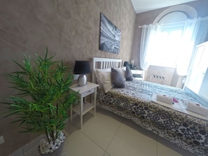Un dormitorio con cama, mesita de noche, lámpara, planta en maceta y ventana con cortinas.