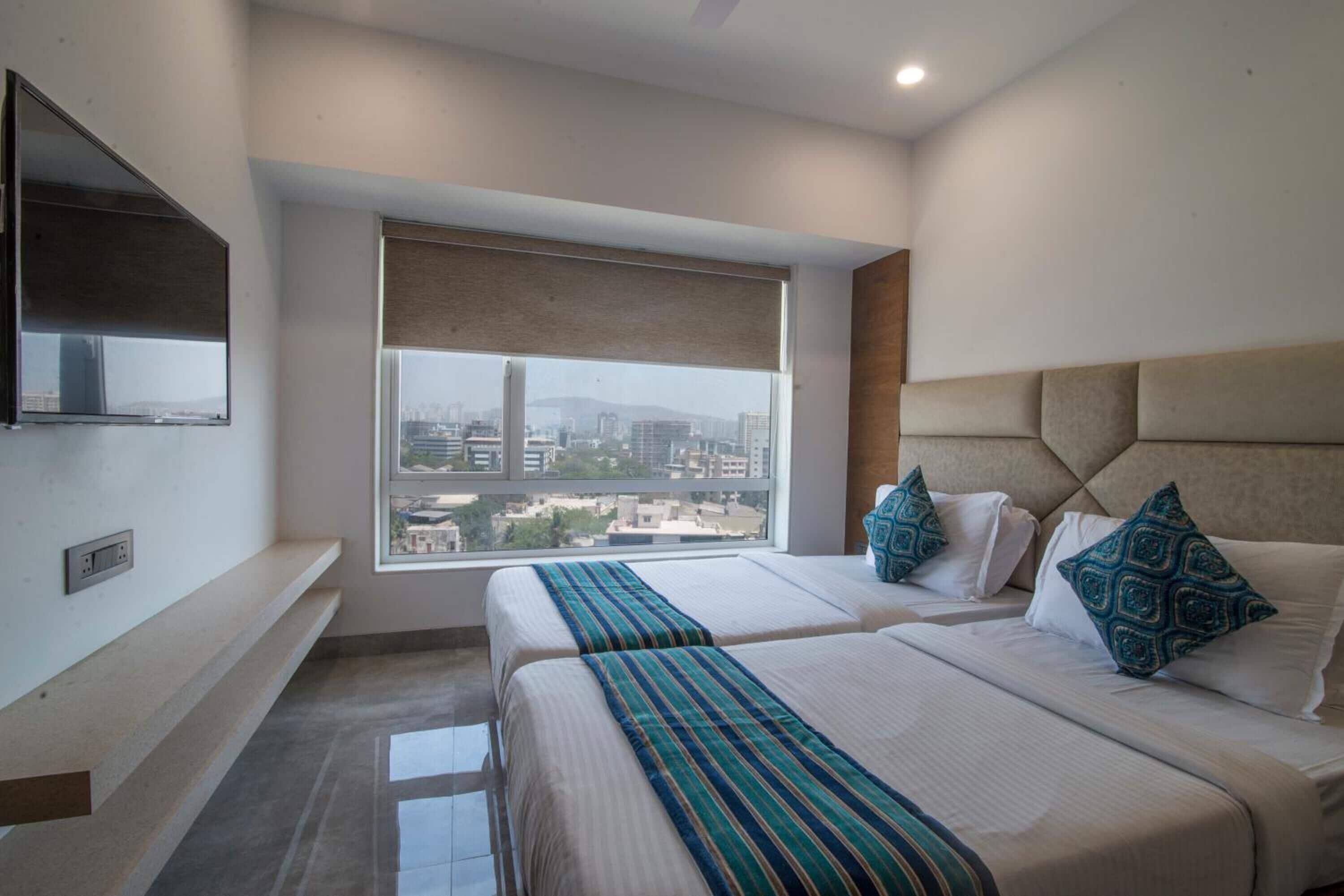 A hotel room with two beds, a large window with a city view, a flat-screen TV mounted on the wall, and a ceiling fan.