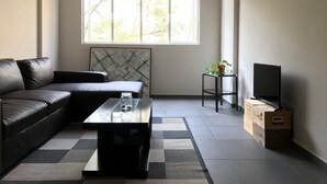 Un soggiorno moderno con un divano in pelle nera, un tavolino da caffè con piano in vetro, un piccolo tavolino laterale con una pianta, un televisore su un supporto in legno e un'ampia finestra con vista sugli alberi.