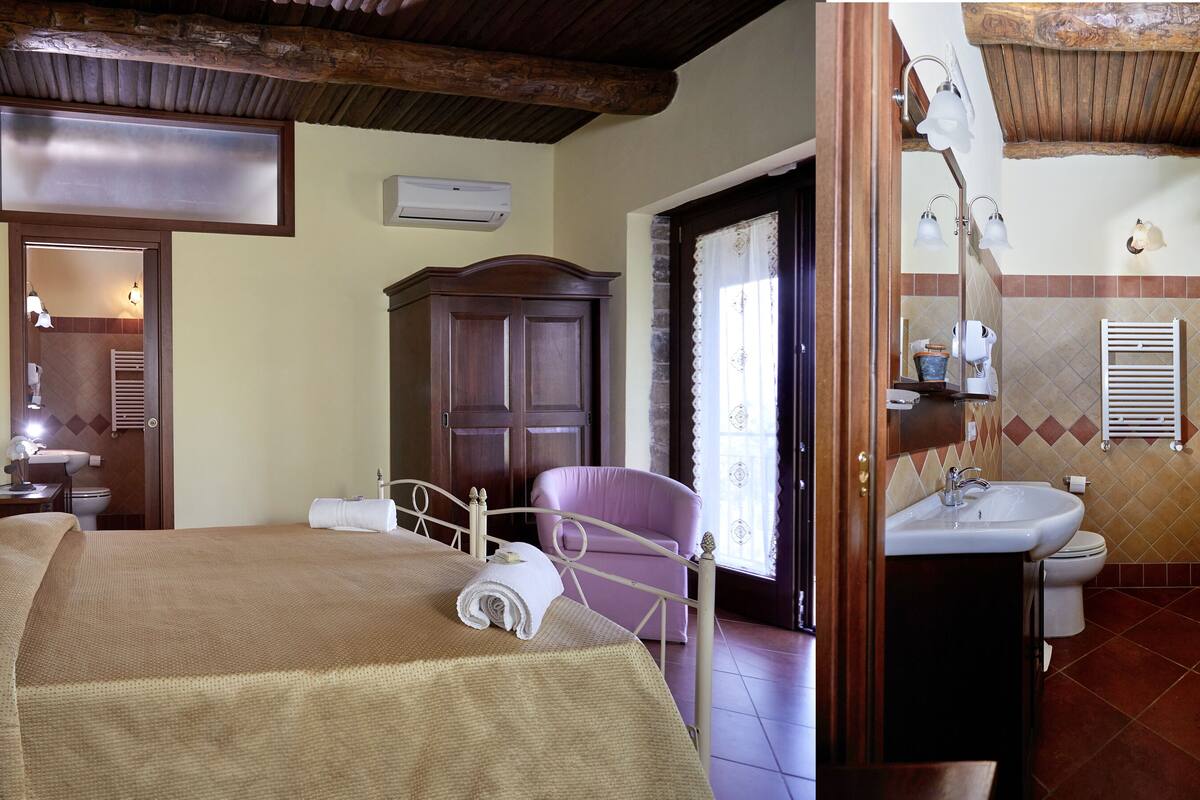 A bedroom with a bed, bedside table, and a chair. There is a bathroom with a sink, toilet, and shower.