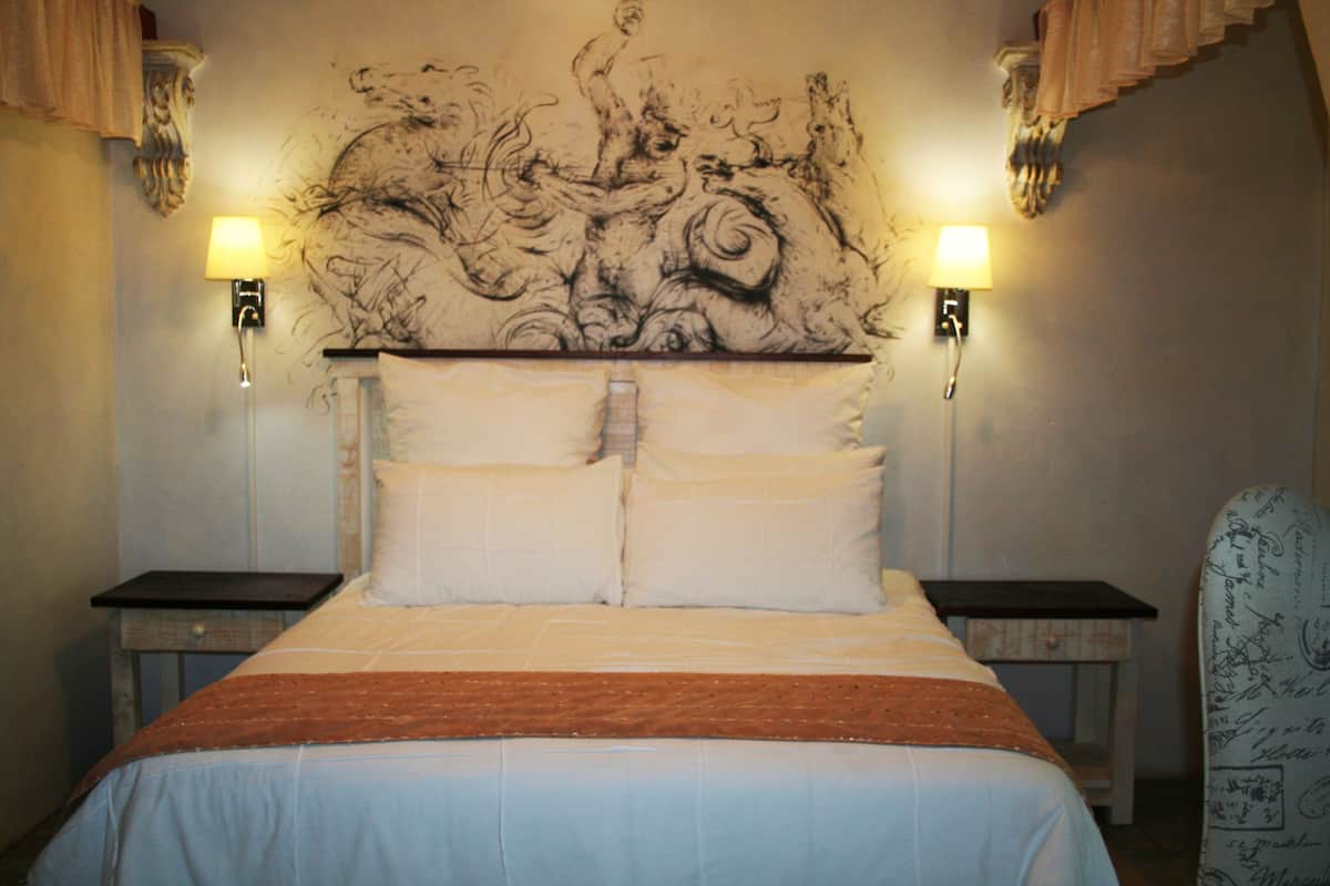 Una habitación de hotel con una cama grande, dos mesitas de noche, una silla y un cabecero decorativo.