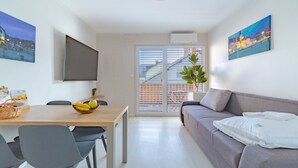 A modern living room with a sofa, a dining table with chairs, a TV mounted on the wall, and a large window with a view of buildings.
