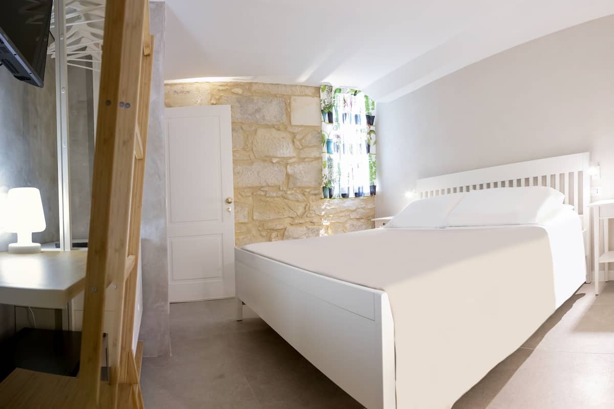 Un dormitorio con pared de piedra, una cama con ropa blanca, una mesita de noche y una ventana con plantas en macetas.
