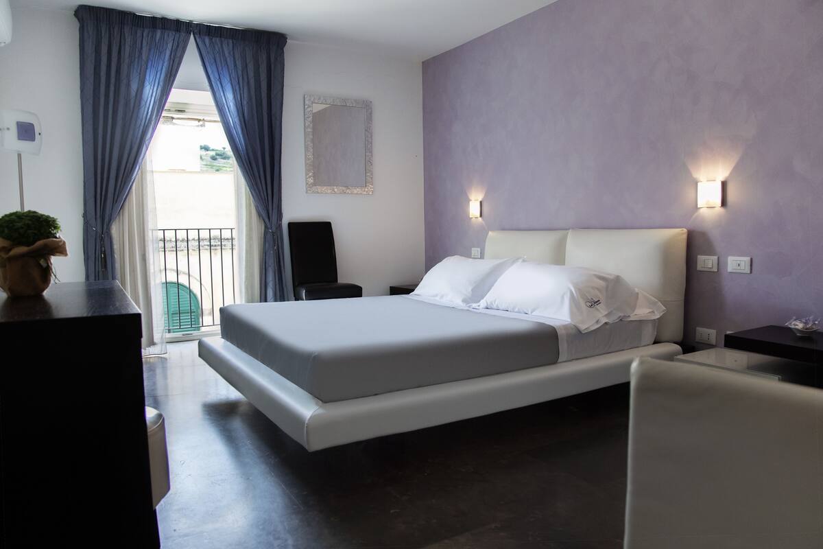 Un dormitorio moderno con una cama grande, una mesita de noche, una silla, una pequeña mesa, un espejo y una ventana con cortinas.