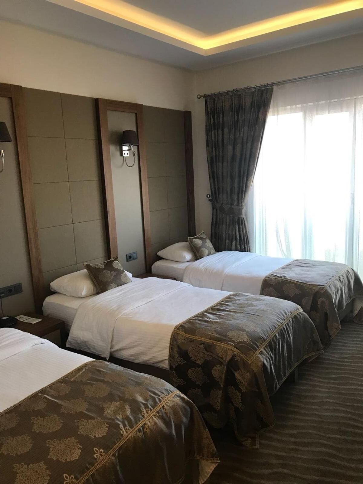 A hotel room with three beds arranged in a row, a large mirror, a wall-mounted lamp, and a window with curtains.