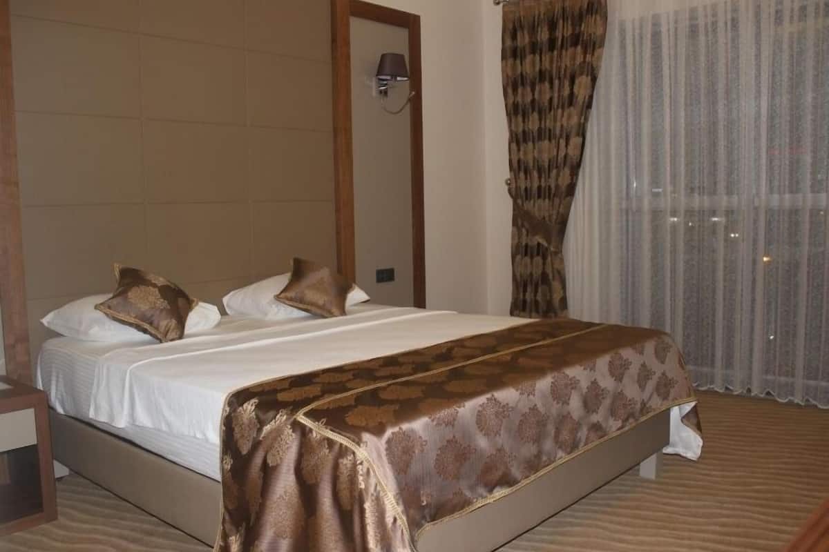 Une chambre d’hôtel avec un grand lit, une tête de lit en bois, une table de chevet et une fenêtre ornée de rideaux légers.
