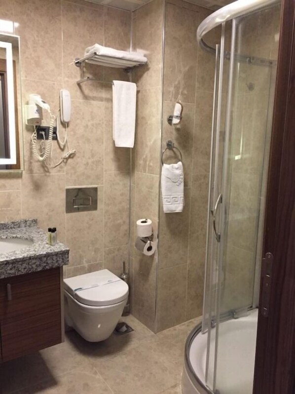 Casa de banho com sanita, lavatório e chuveiro. Há toalhas e um secador de cabelo na parede.