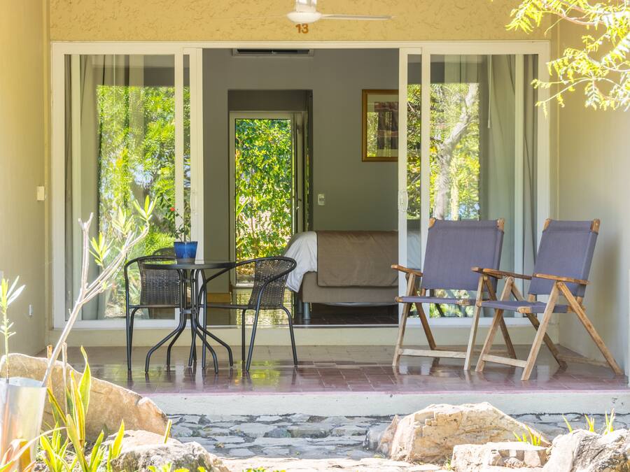 Un espacio exterior techado con una mesa y dos sillas, una puerta que da a una habitación, y vistas al exterior con vegetación.