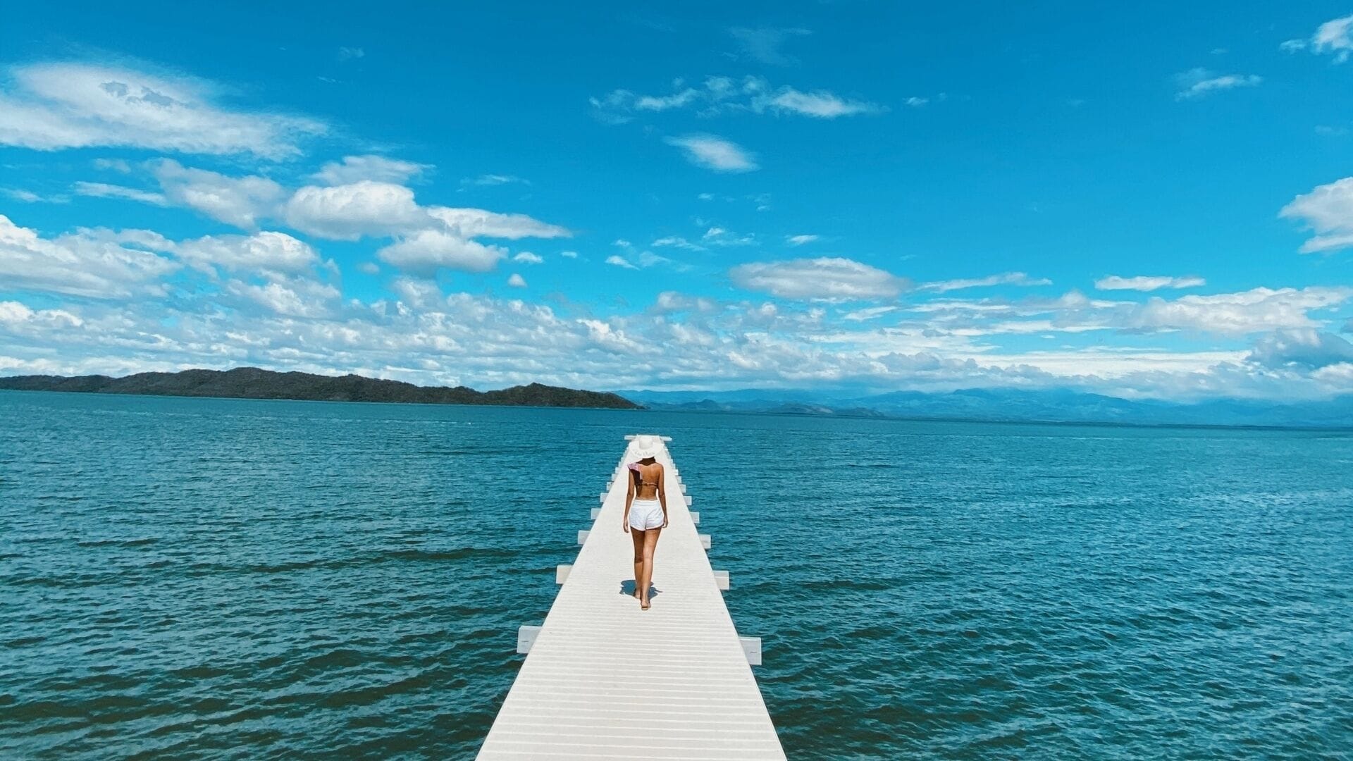 A woman walking on a white pier extending into a calm sea, with mountains in the background under a clear sky.