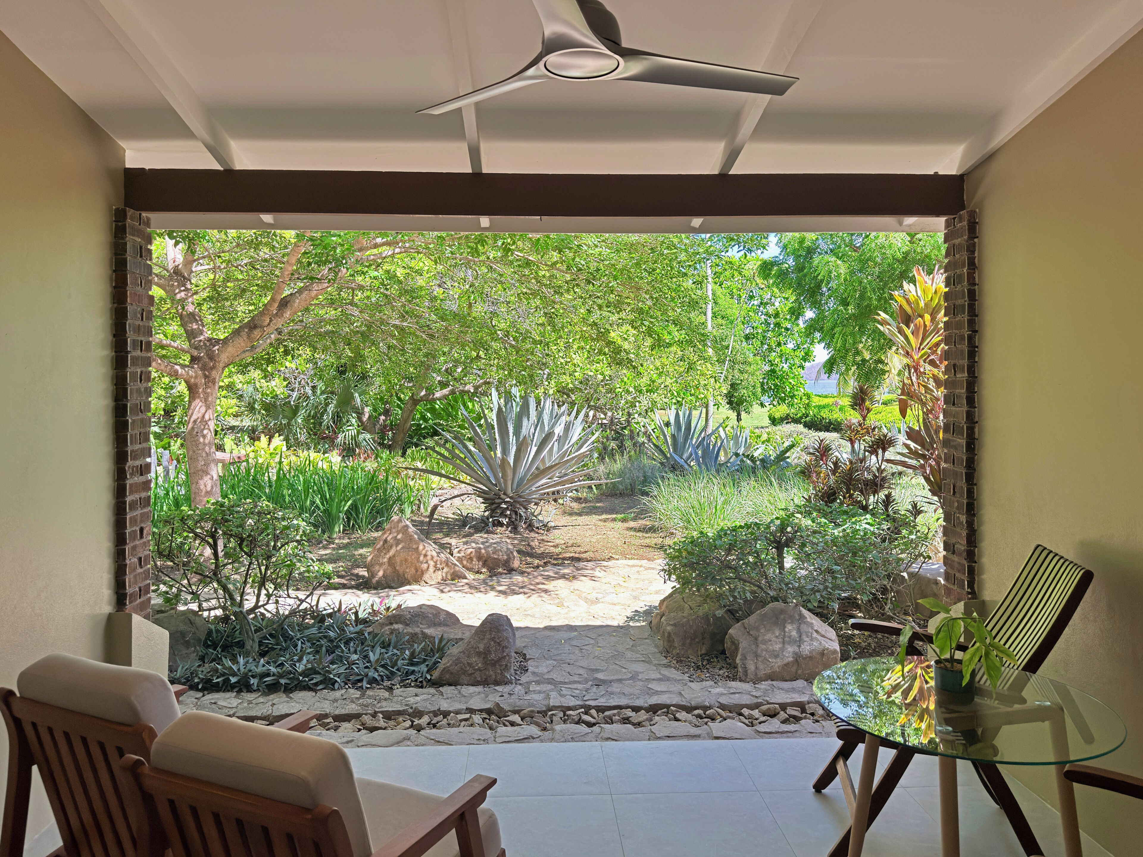 A room with a glass table, a ceiling fan, and a view of a garden with rocks and plants.