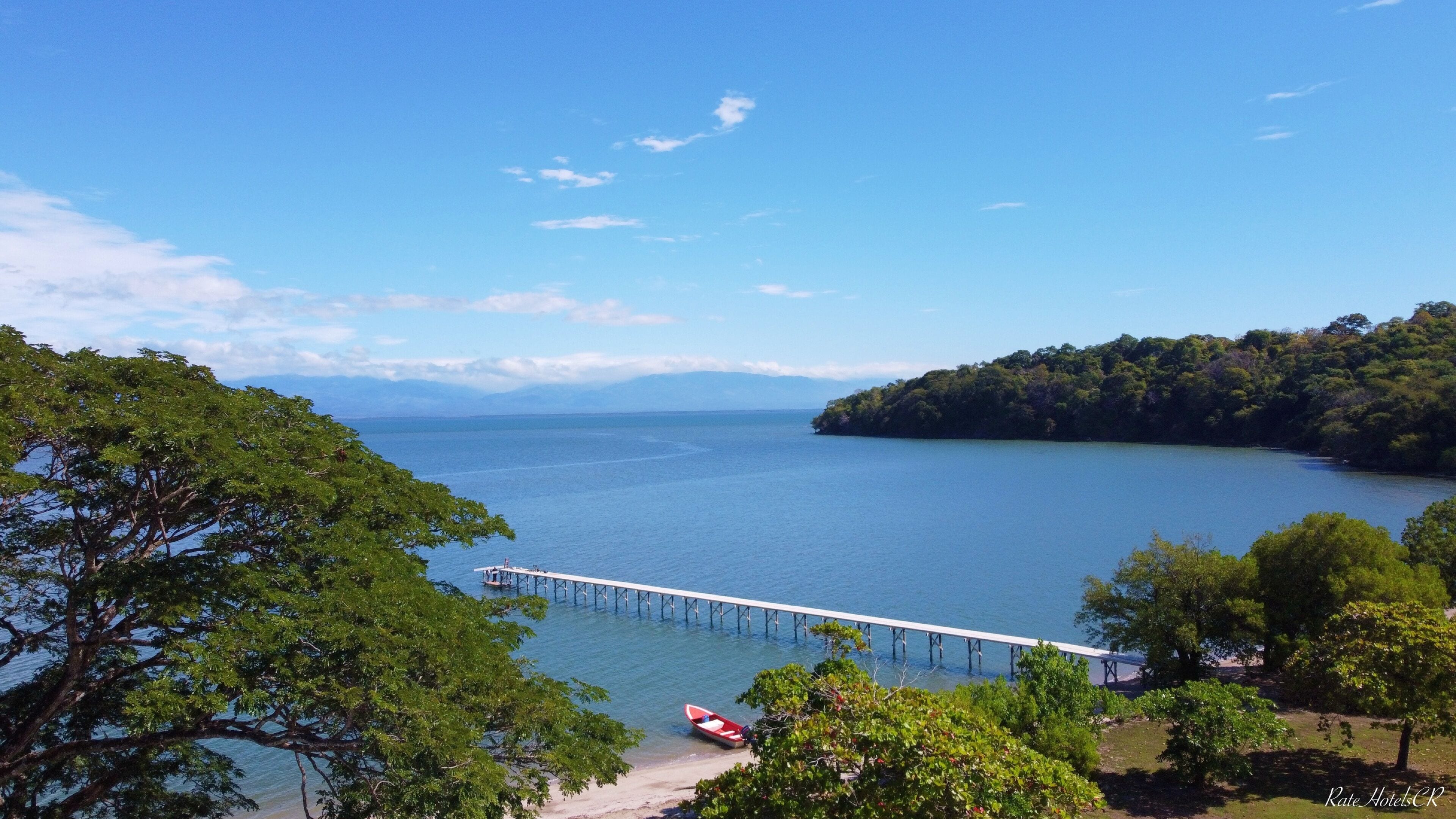 Vista costeira com um cais que se estende sobre a água, rodeado por vegetação exuberante e um céu claro.