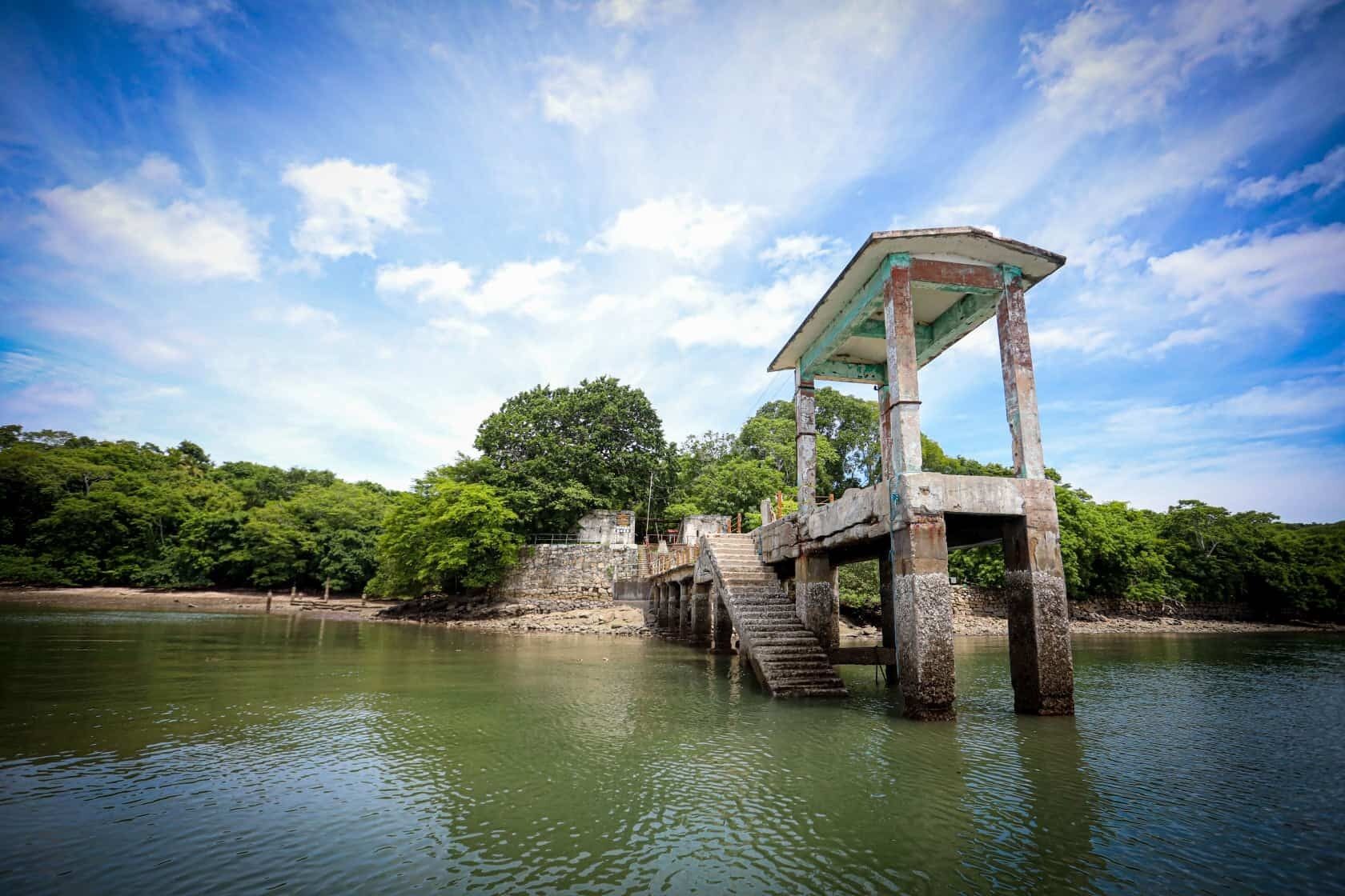 A concrete structure with a roof and stairs leading to a platform over water, surrounded by greenery.