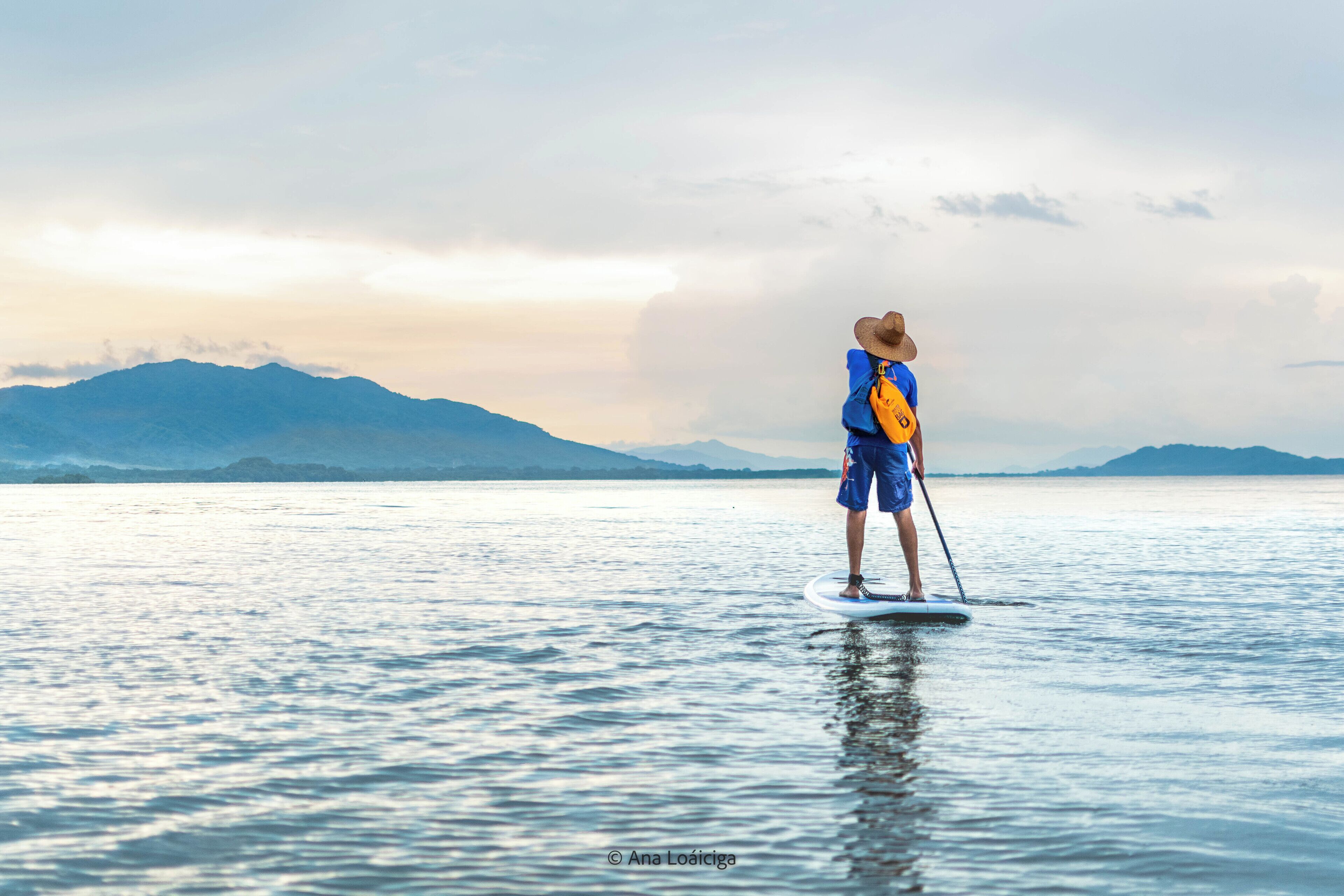 Eine Person steht auf einem Paddleboard in ruhigem Wasser, im Hintergrund sind Berge zu sehen.