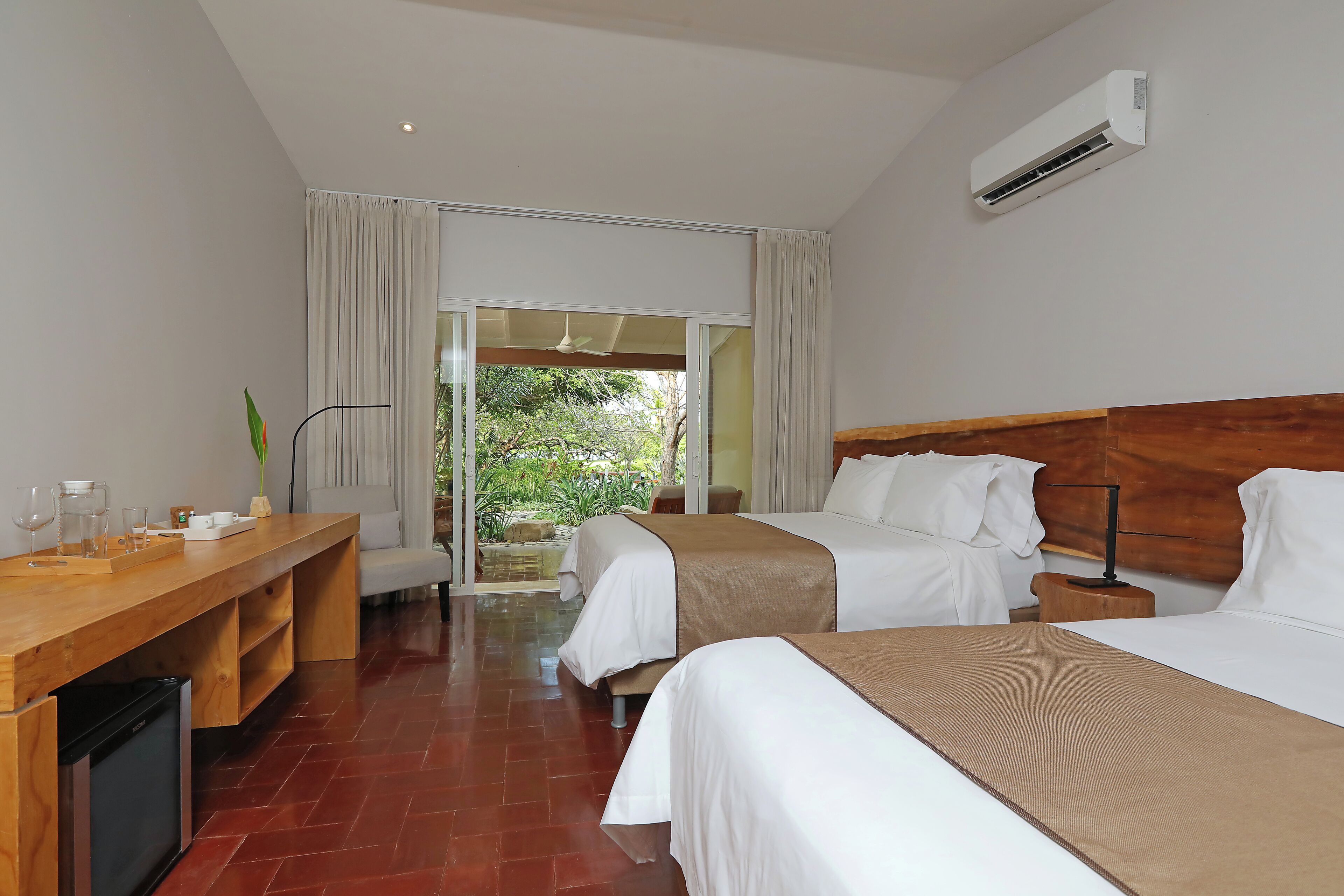 A hotel room with two beds, a wooden desk, a small fridge, and a view of greenery outside.