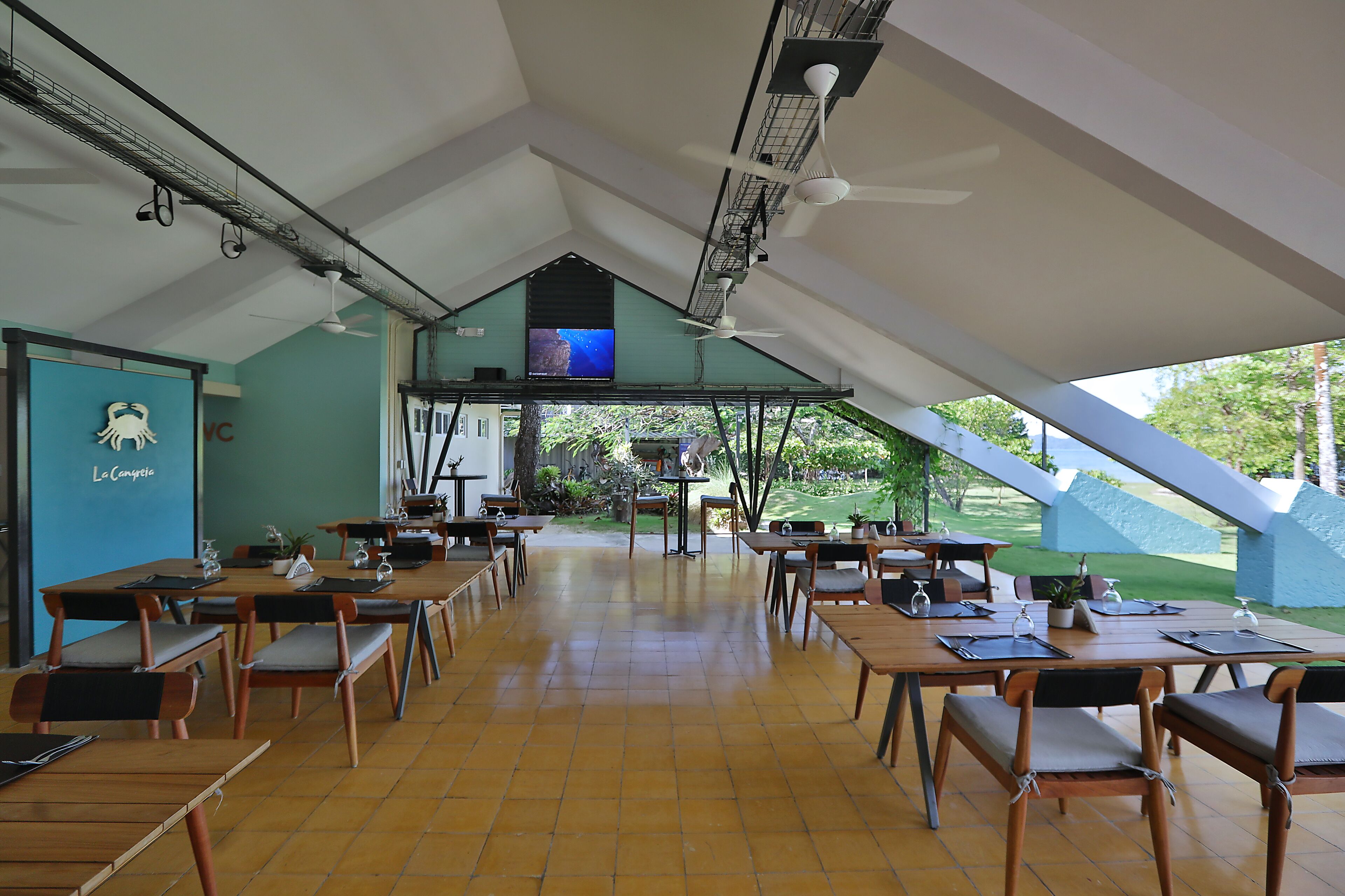 Un vaste espace dédié à la salle à manger, doté de hauts plafonds et baigné de lumière naturelle, comprenant plusieurs tables et chaises, un écran de télévision et une vue sur l’extérieur.