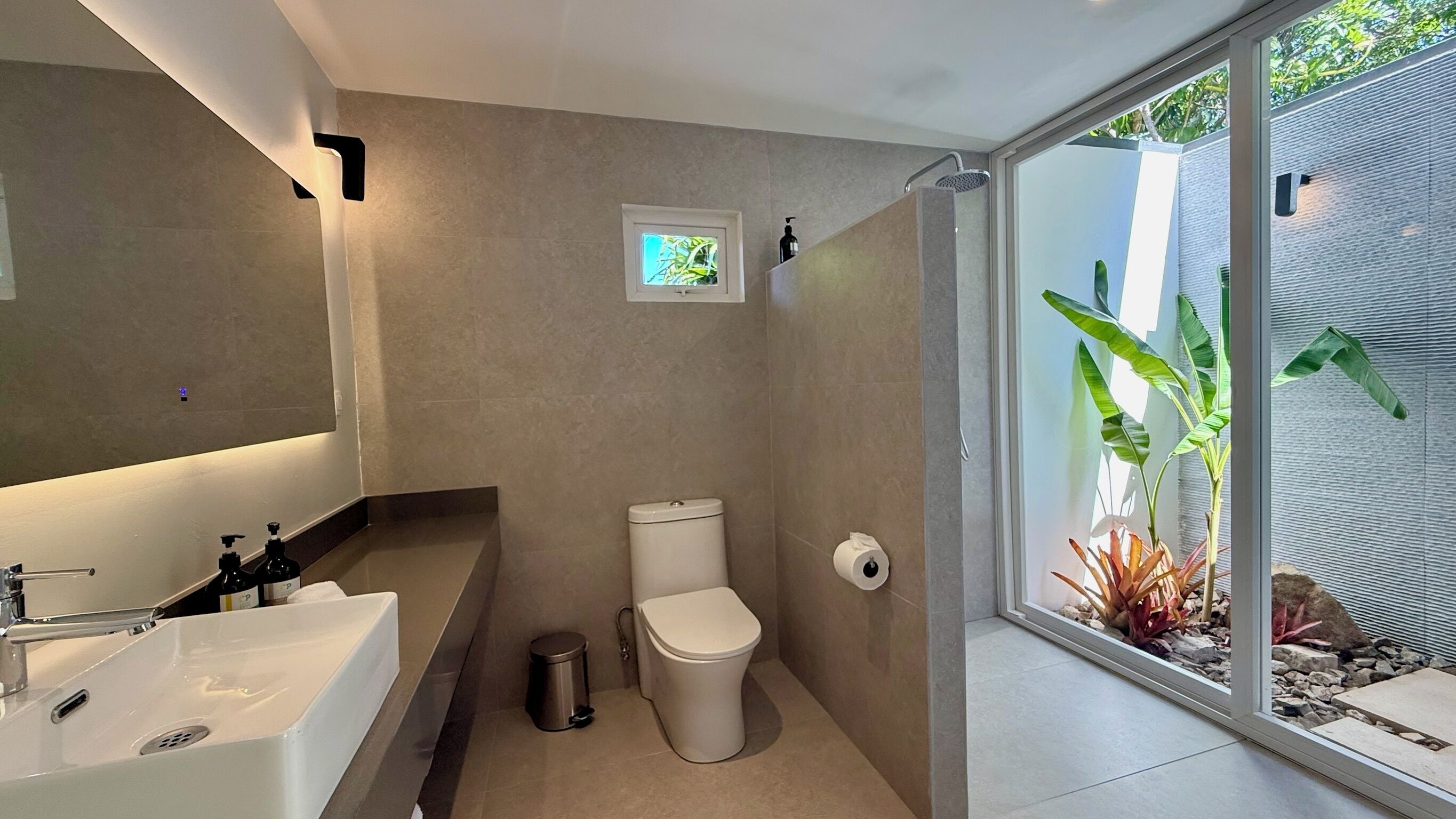 Un baño moderno con lavamanos, inodoro y ducha blancos. Hay una ventana pequeña con vistas a plantas afuera.
