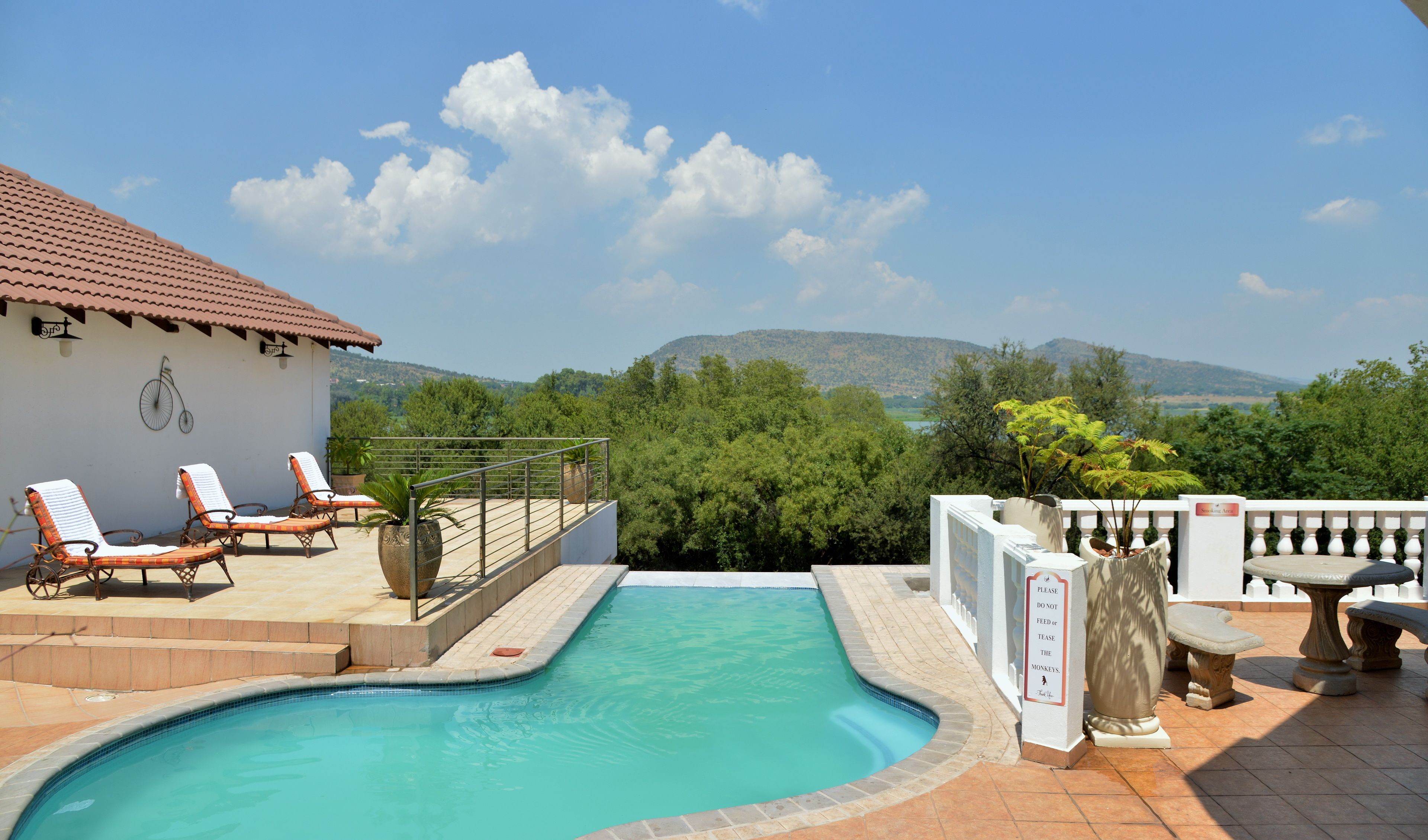 A swimming pool with lounge chairs, a sign prohibiting feeding monkeys, and a view of hills.