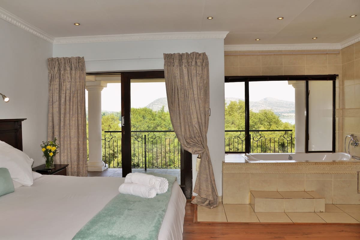 Een slaapkamer met een groot bed, uitzicht op groen en bergen, een balkon met een zwarte railing, en een aparte badkamer met een bad.
