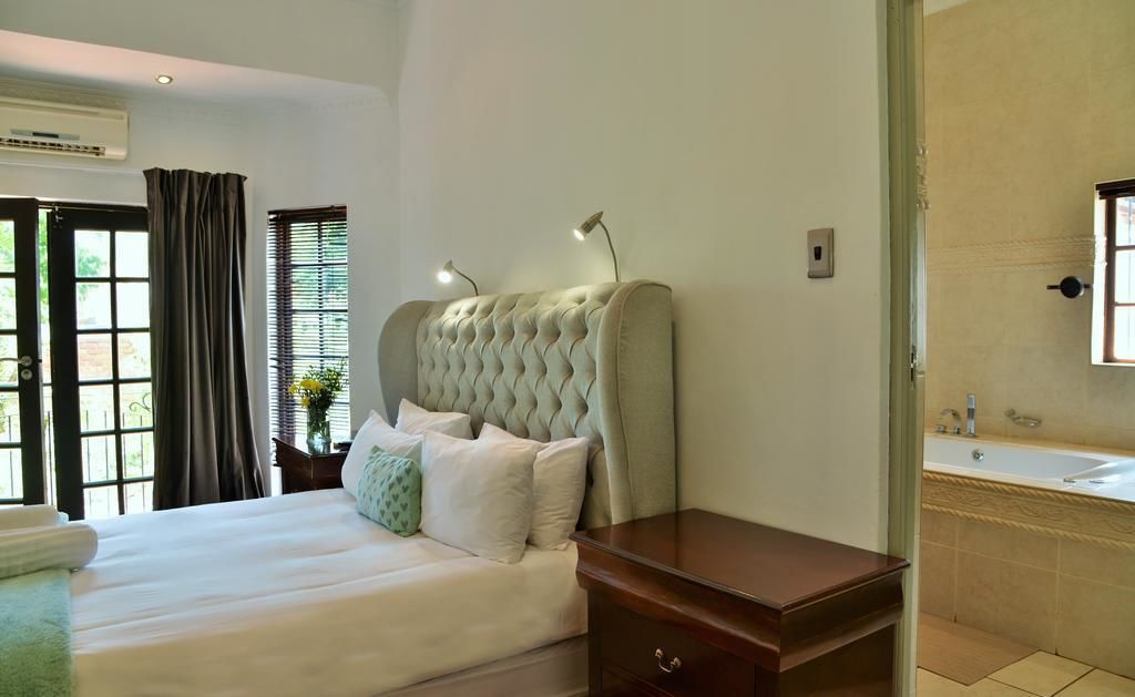 A bedroom with a large bed, a bedside table, a nightstand, a window with blinds, and a bathroom with a sink and mirror.
