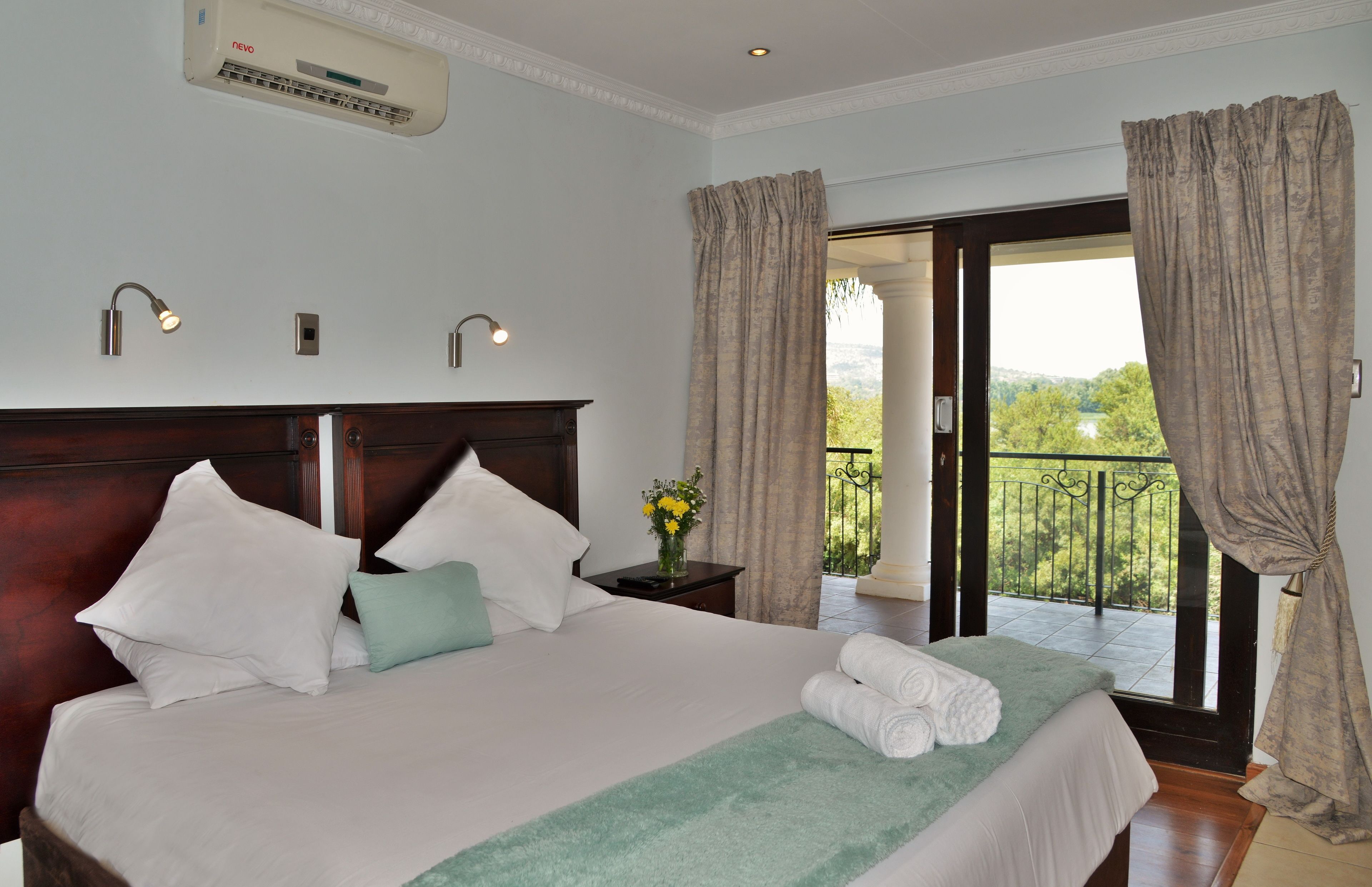 A hotel room with a large bed, a nightstand with a vase of flowers, and a view of a balcony with a railing.