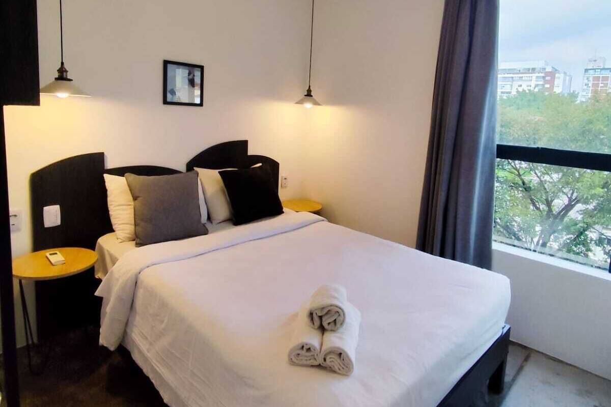 Habitación de hotel con cama, mesitas de noche, lámpara, un cuadro en la pared y ventana con vistas a edificios y árboles.
