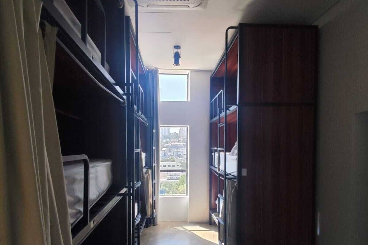 Un pasillo estrecho con literas a ambos lados, una ventana al final y un equipo de aire acondicionado en el techo.