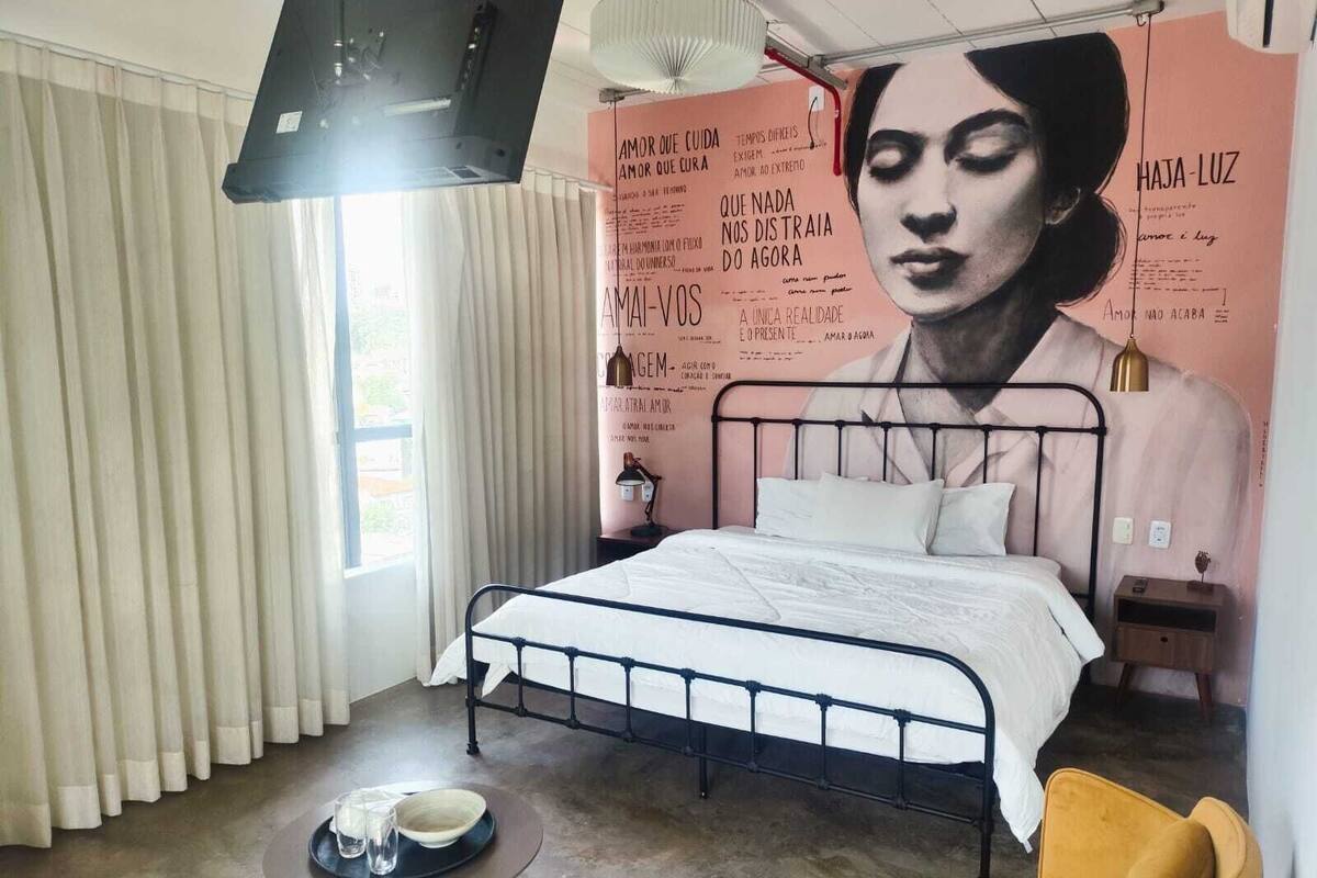 Un dormitorio con cama, mesita de noche, silla, una pequeña mesa con vasos, y una pared con texto y parte de la imagen de una persona.
