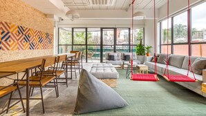 Un espacio de oficina moderno con una larga mesa de madera, múltiples sillas, un sillón de saco gris, un sofá gris con cojines rojos y ventanas amplias que ofrecen vistas de la ciudad.