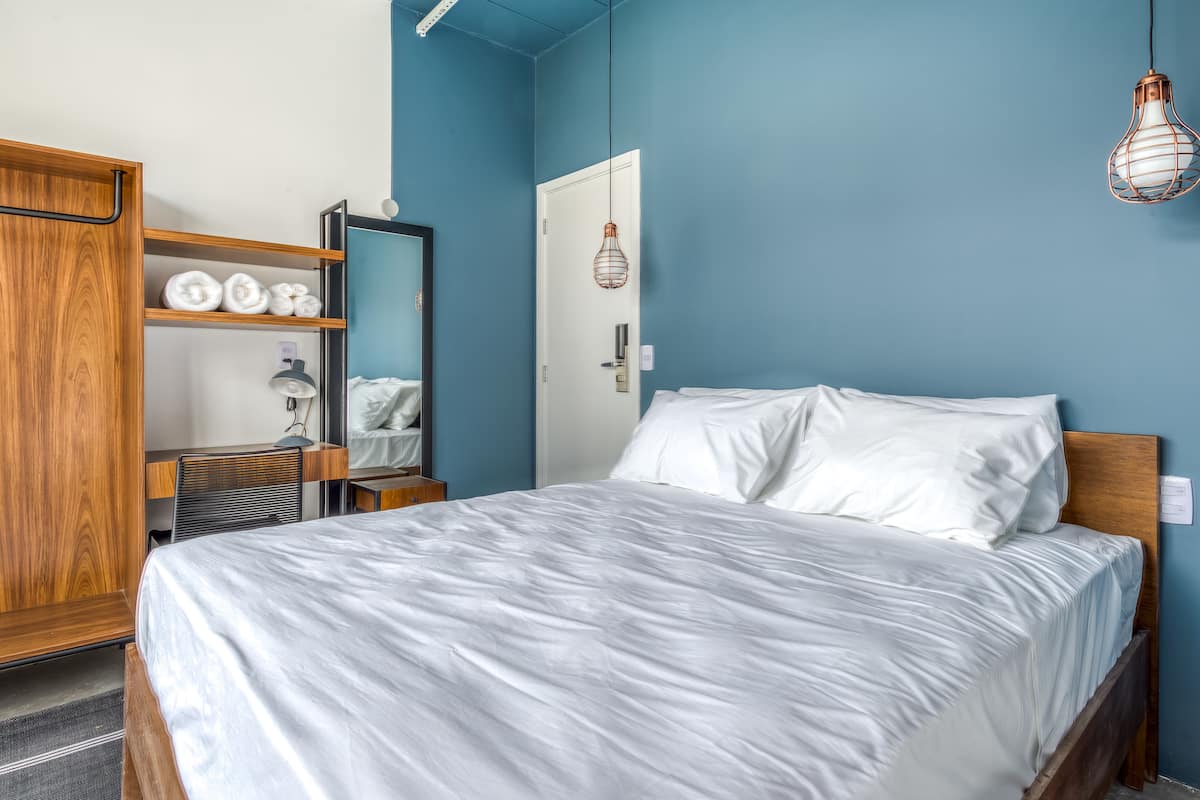 Una cama bien hecha con sábanas blancas, un armario de madera con repisas, una puerta con lámpara y una pared azul.
