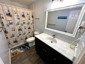 A bathroom with a shower curtain featuring lighthouse prints, a white toilet, a sink with a mirror above it, and a hairdryer on the wall.