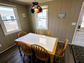 A dining area with a square table, four wooden chairs, a colorful hanging light fixture, and a 'FLIP FLOP ZONE' sign on the wall.