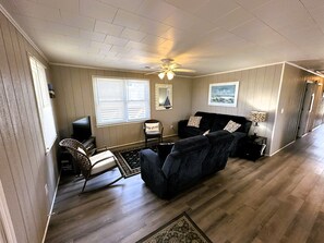 Ein Wohnzimmer mit einer schwarzen Couch, einem Schaukelstuhl, einem Deckenventilator und einem Fenster mit Jalousien.