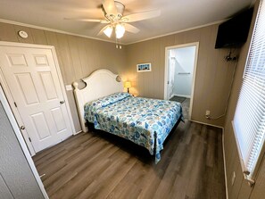 A bedroom with a bed, a ceiling fan, a door, a window with blinds, and a wall-mounted television.