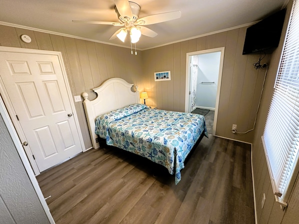 A bedroom with a bed, a ceiling fan, a door, a window with blinds, and a wall-mounted television.