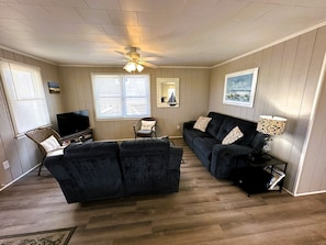 A living room with a dark-colored sofa, a television, a ceiling fan, and a window with blinds.