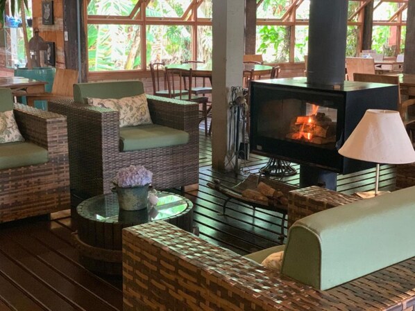 A cozy lounge area with wicker furniture, a fireplace, and large windows offering a view of greenery.