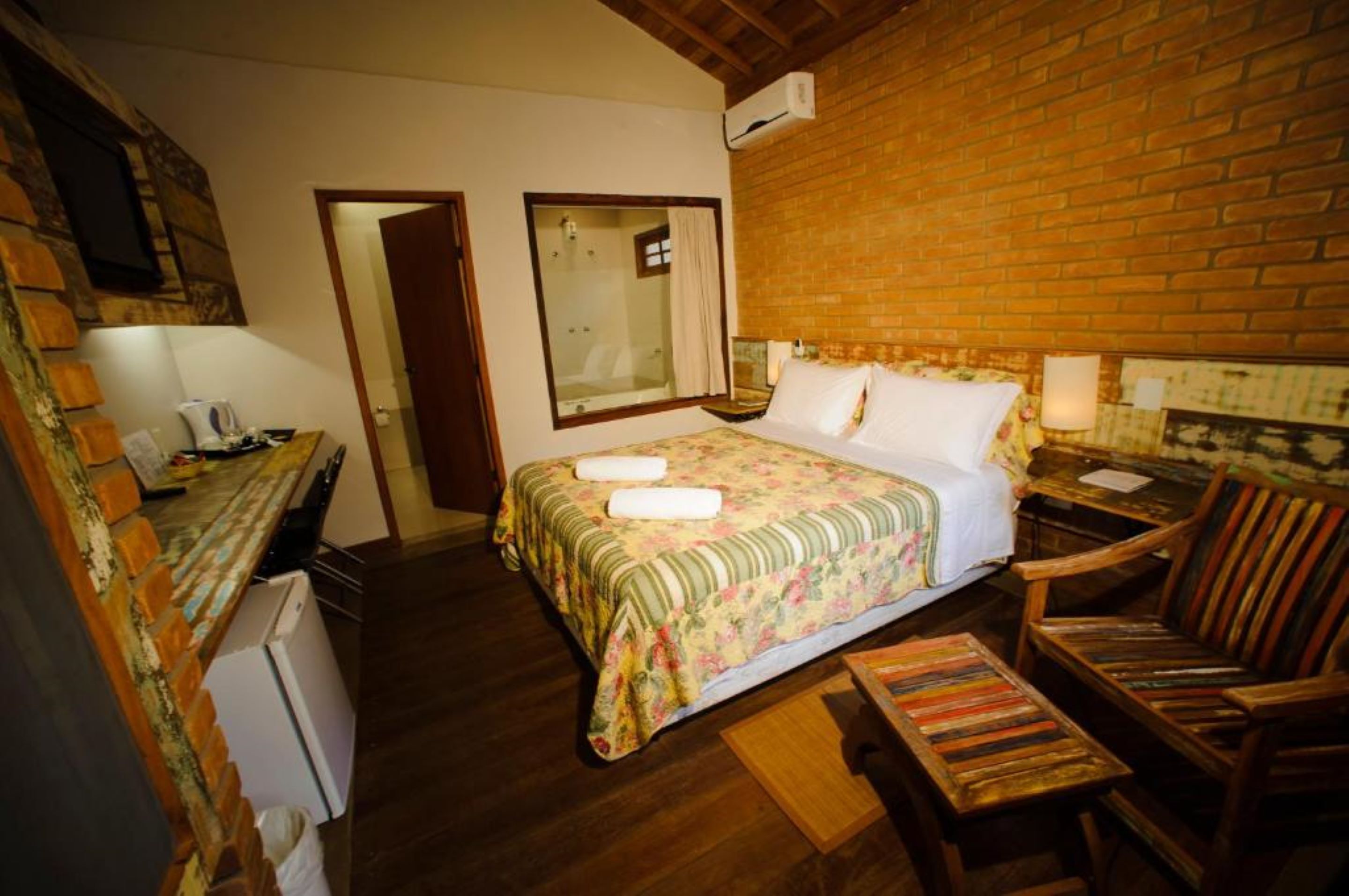 Um quarto com cama, uma cadeira de madeira, uma mesinha, um espelho e uma parede de tijolos.
