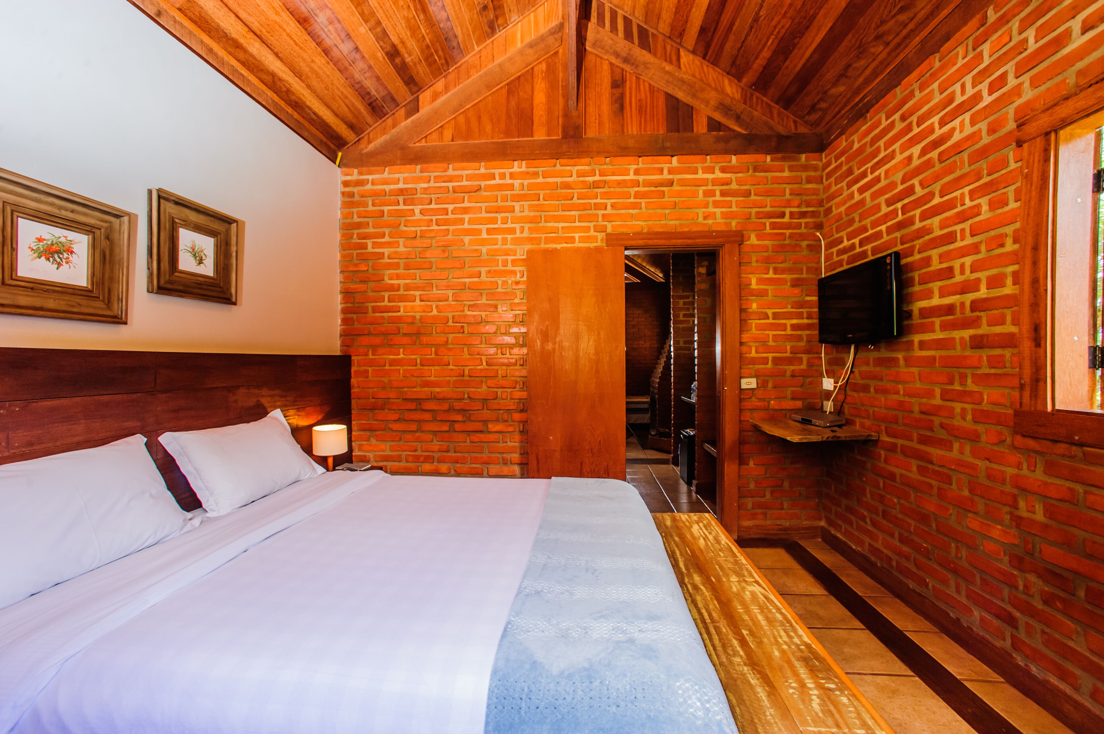 A bedroom with a brick wall, wooden ceiling, a bed with white linens, a TV mounted on the wall, and a door leading to another room.