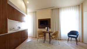 Cocina moderna con armarios de madera, estufa integrada y televisor de pantalla plana montado en la pared. Hay una mesa redonda con dos sillas y una silla azul en una sala de estar con un amplio ventanal y cortinas.