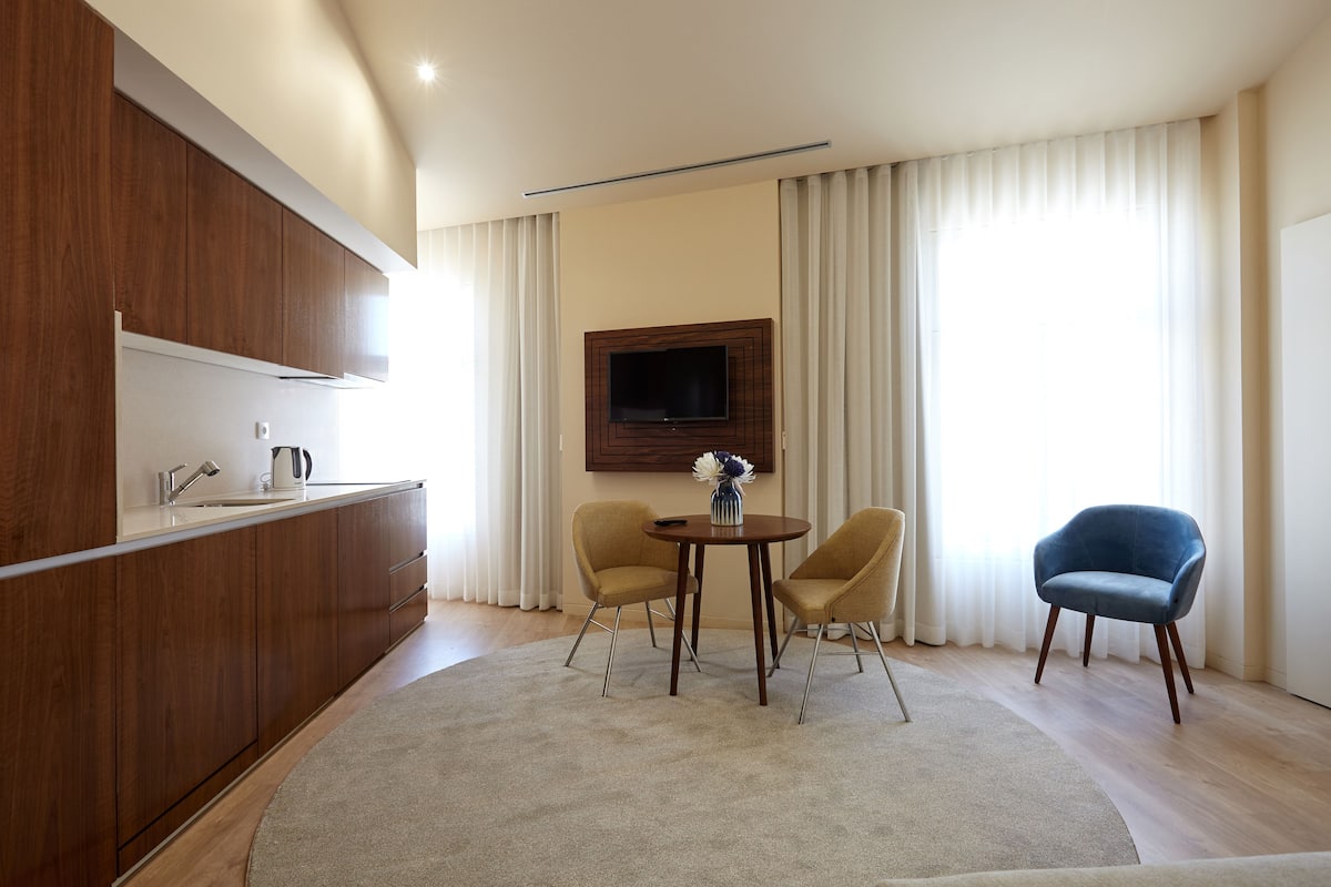 Eine moderne Küche mit Holzausstattung, integriertem Herd und einem flachbildfernseher an der Wand. Ein runder Tisch mit zwei Stühlen und ein blauer Sessel befinden sich in einem Wohnbereich mit großem Fenster und Vorhängen.