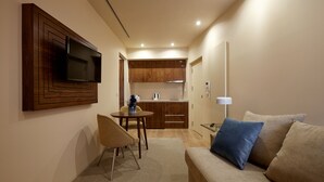 Un espacio habitable compacto con kitchenette, televisor de pantalla plana montado en la pared, sofá con cojines y una pequeña mesa de comedor.