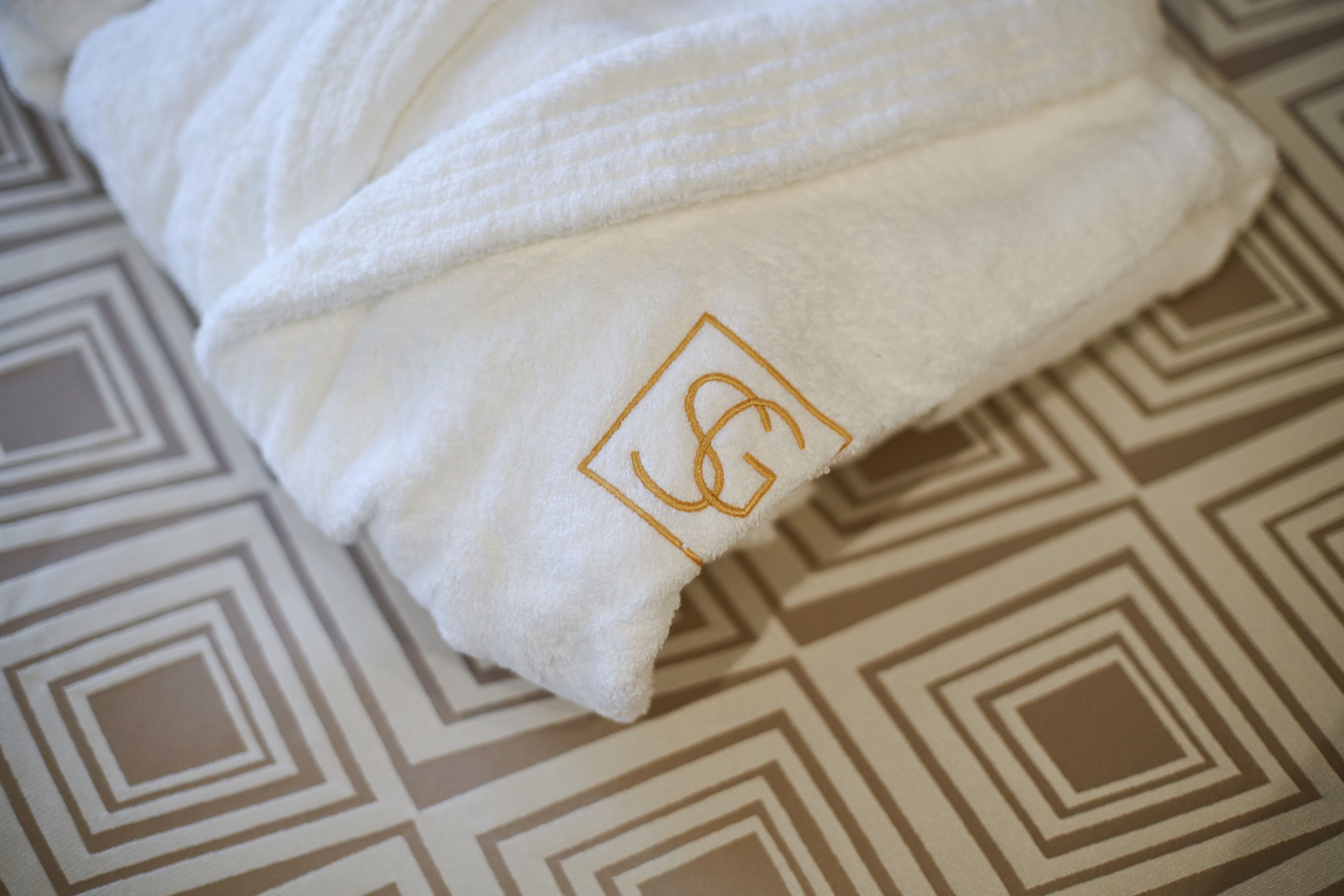 Una toalla blanca con un logotipo bordado en dorado sobre un suelo estampado.